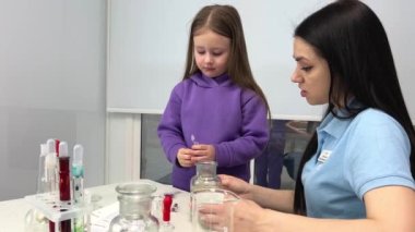 laboratory of chemical experiments for children little girl pours sequins from test tube special liquid for bubbles nearby the laboratory assistant helps shows poured lot Lilac suit Blue t-shirt