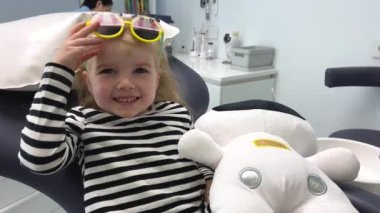 a little cheerful girl in dental chair with a bruise on her forehead puts on sunglasses they fall down, she corrects them, sits with a toy in the background, a nurse