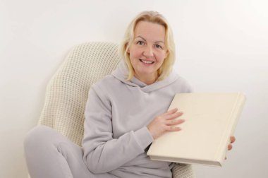 a woman with a white album in her hands on sofa with a plaid in an armchair in a tracksuit Memories looks into the frame
