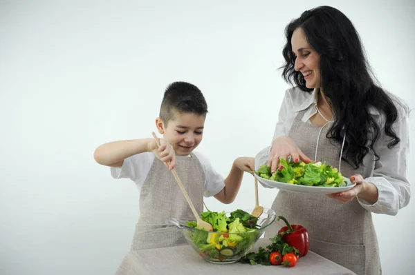 mom and son cooking fresh vegetable salad boy happy stirs food with two wooden spoons mom yes sprinkles lettuce leaves smiling matching clothes apron white background studio space for text grocery