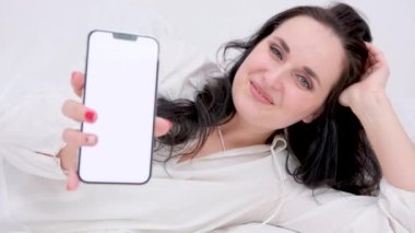 beautiful brunette holds phone with white screen close-up smiles showing ad laughs with finger shows at phone thumbs up really likes lies on side supports head with hand advertising space for text