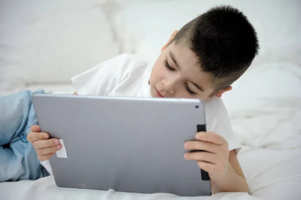 little boy 7 years old lies on bed in hands with tablet online learning interesting-movie mobile games applications I am alone at home spend time on the Internet social networks children and computer