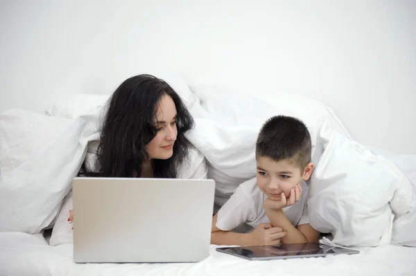 mom and son 7 years old lying under blanket on white sheets boy looking at tablet playing game mom turned to face him love for children care online learning english course learning technology getting