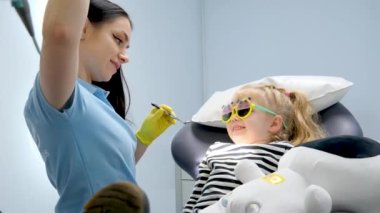 pediatric dentistry young beautiful doctor in yellow gloves turns on light showing girl the tools with which to treat teeth caries color directs to girls face on sunglasses in hands a soft toy