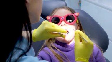 Pediatrik diş hekimliği. Kapalı çenesi olan küçük bir kız. Diş hekimi, dişçi kızın diş macunu takması için kırmızı güneş gözlüğü takıp takmadığını kontrol ediyor.