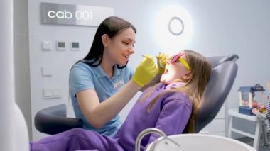 Pediatrik diş hekimliği. Kapalı çenesi olan küçük bir kız. Diş hekimi, dişçi kızın diş macunu takması için kırmızı güneş gözlüğü takıp takmadığını kontrol ediyor.