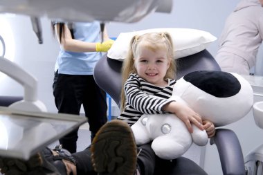 close-up of little girl with soft toy in hands shows tongue and looks shy to sides nurse walks to right to left the camera is approaching striped blouse dental chair hospital social assistance