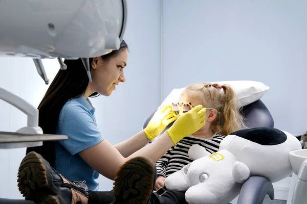 dental chair doctor in mask with yellow gloves with tool for dental treatment in hand sits near little girl who tries on sunglasses doctor waits for child to be ready for treatment dental filling
