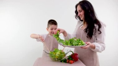 Anne ve oğlu taze sebze salatası pişiriyor. Salatacı çocuk, iki tahta kaşıkla yemek pişiriyor. Anne, marul yaprakları serpiştiriyor.