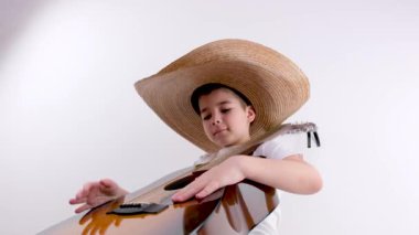 Küçük bir çocuk sandalyeye oturur ve elinde bir gitar tutar. Şapkalı bir çocuk beyaz arka planda gitar çalar. Rahatlar, müzik aleti çalar.