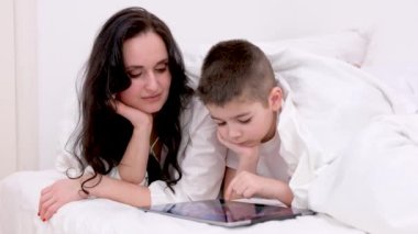 mom and son 7 years old lying under blanket on white sheets boy looking at tablet playing game mom turned to face him love for children care online learning english course learning technology getting