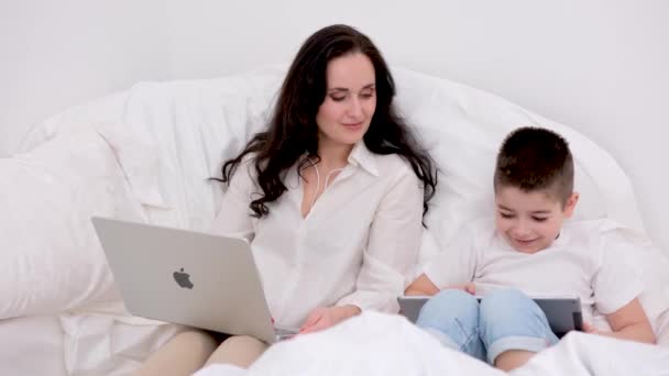 お母さんと息子は彼らの手の中に白いベッドに座っていますアップル技術のラップトップMacbookタブレットIpadと携帯電話Iphone 13プロマックスお互いの笑顔を見て 最新の技術を使用していますウクライナ2023 — ストック動画