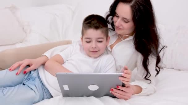 妈妈和儿子舒舒服服地躺在床上 在一家漂亮的白色宾馆里玩着一款叫做 的平板电脑游戏 — 图库视频影像