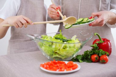 Professional female cook adding fresh cucumber slices in glass bowl with salad. mother and child preparing vegetable salad close-up boys hands help mother to add cucumbers glass plate