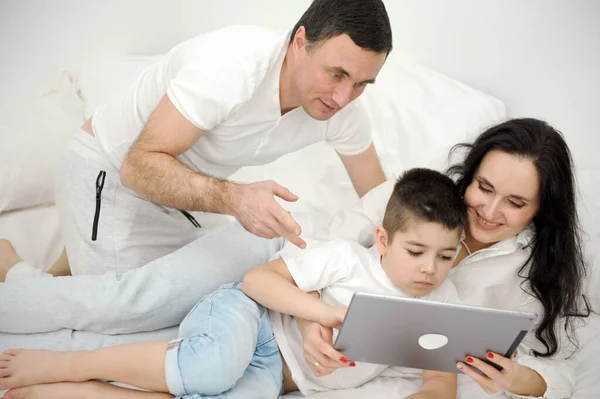 people, family and technology concept mother father and little boy with tablet pc computer in bed at home or hotel room. Happy family spend time together lying under white blanket on bed