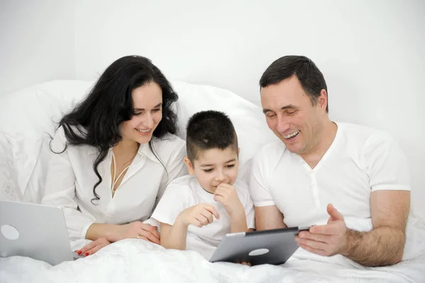 people, family and technology concept mother father and little boy with tablet pc computer in bed at home or hotel room. Happy family spend time together lying under white blanket on bed