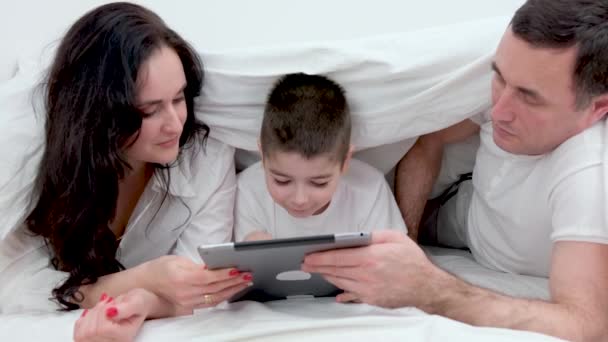 家庭和科技概念妈妈爸爸和小男孩在家里或宾馆的床上带着平板电脑 快乐的家人一起躺在白色毯子下躺在床上 — 图库视频影像