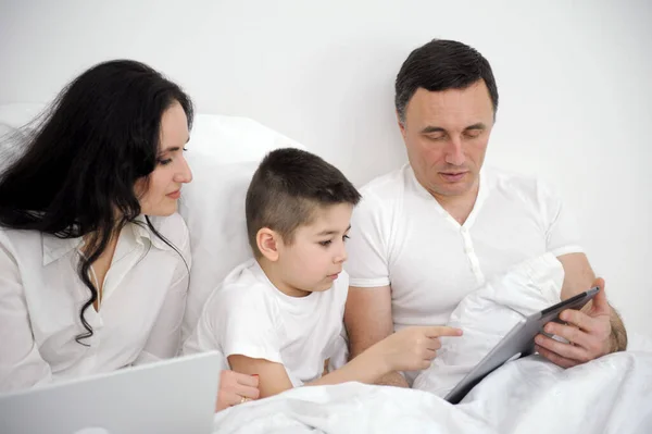 banner latest technology family husband wife and child sitting watching movie on laptop on bed relaxing together white clothes chatting online with relatives mom dad and son laughing smiling rejoicing