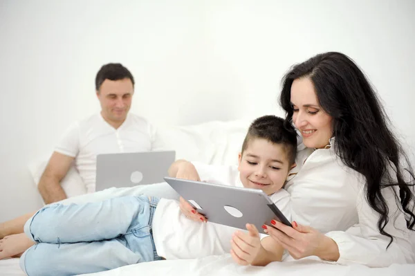 latest technology gadgets mom and son playing on tablet father sitting on bed working on laptop remote work freelancer vacation family combine work and spend time with family together