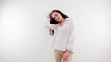 beautiful brunette on a white background straightens her hair beige leggings white shirt gentle smile beauty cleanliness long thin arms beautiful figure young body 30-40 years old woman girl space ad