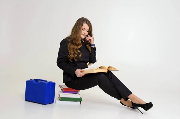 need knowledge woman sitting on stack of books on white background in studio reading thick large vintage book smart watch suitcase tools medical doctor in black high-heeled suit professor teacher bag