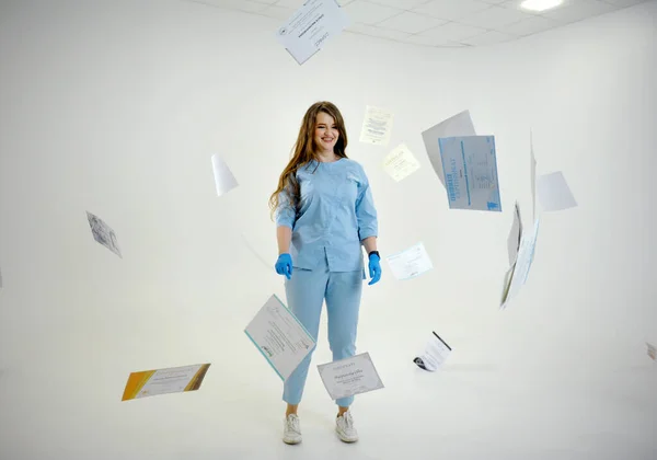 female doctor in blue medical suit and hygienic gloves stands in center of white room certificates of graduation from medical university flying around training dental college education passing years