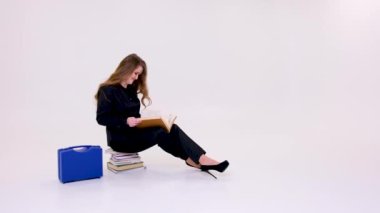 need knowledge woman sitting on stack of books on white background in studio reading thick large vintage book smart watch suitcase tools medical doctor in black high-heeled suit professor teacher bag