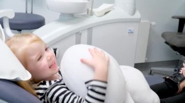 Dişçi koltuğundaki küçük kız oyuncağıyla oynarken eğleniyor gülümseyen çizgili bluzla konuşuyor pediatrik dişçide çocukları eğlendirmek için son teknoloji.