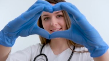 Doctor hands in gloves in the heart shape against the background of his body face and medical gown. closeup. Female hand in blue medical gloves show heart sign. High quality photo
