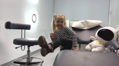Diş hekimine git küçük kız çocuğu neşeli bir gülümsemeyle dişçi sandalyesine tırmanıyor küçük çocukların küçük bacakları üzerinde büyük ayaklı yumuşak oyuncakları kapıyor erken yaşta diş tedavisi görüyor.