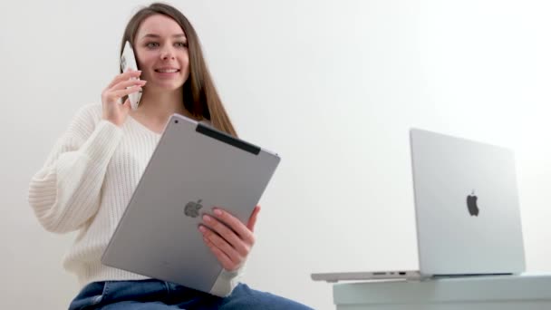 最新の技術Apple MacbookのIpadと携帯電話白いジャケットの女の子の手の中に携帯電話の仕事で話す笑顔便利な快適なオフィスの白い背景スペーステキスト広告のための — ストック動画
