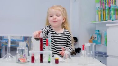 Çizgili kazaklı bir kız, kimya laboratuvarında çocuklara fizik laboratuarı asistanı öğretmek için kırmızı boyalı bir test tüpü seçiyor.