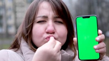 Yetişkin bir kadın, yağmurda, elinde telefon, yeşil ekran kromatonuyla, nasıl bir öpücükle reklamları sevdiğini gösteriyor. Gülümsüyor, üç parmağını öpüyor ve yana doğru yayıyor.