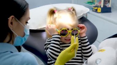 close-up face of a little girl and doctor in a mask yellow hygienic gloves dentist tools pediatric dentistry sunglasses with teddy bear eyes girl treats teeth shows doctor painless treatment