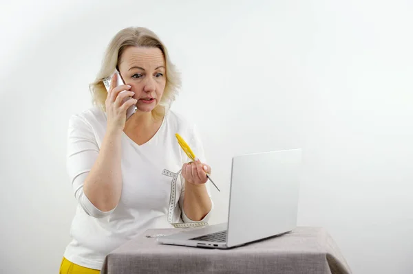 serious dissatisfied woman talking on phone looks into a laptop in her hands scissors and a meter weight loss diet tired of communicating with a doctor nutritionist middle age white background