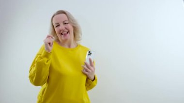 Orta yaşlı yetişkin bir kadın stüdyoda beyaz ekranı olan bir telefonun elinde pozitif duyguların reklamını yapmak için dans ediyor.