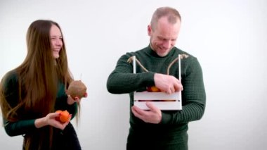 man and woman White wooden box hold fruits apples tangerines persimmon Coconut young girl smiling laughs man put basket around neck looks concept healthy nutrition vitamins benefits good mood fun