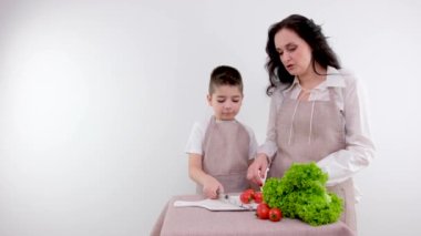 Annesi küçük oğluna kiraz domateslerinin nasıl kesileceğini öğretiyor. Çocuk bir peynir bıçağı alıyor ve beyaz porselen tahta üzerinde domates kesmeye çalışıyor. Özel mutfak önlükleri.
