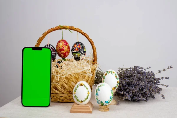 easter composition with phone with white chroma key screen for ad basket of eggs with embroidery eggs painted lavender flowers wooden coasters on a white background Easter holiday
