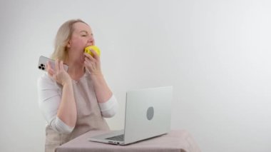housewife cooks food looks at recipes on computer on internet and talks on phone and eats apple on white background space for text cooking cooking delicious food diet nutritionist communicating