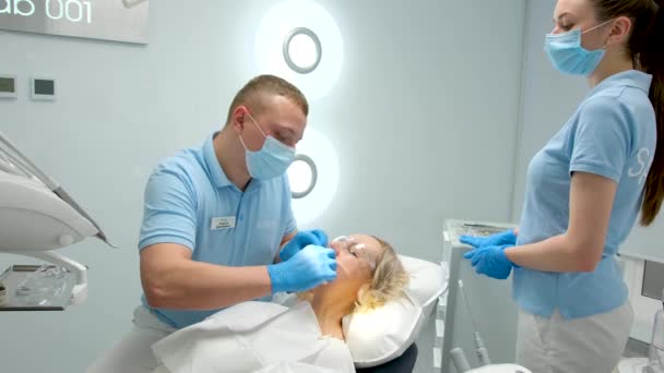 牙科诊所医生在牙科诊所行医前把橡胶坝牵引器放进嘴里清洁牙齿美白充填假肢根管治疗医生和身穿蓝色口罩的护士 — 图库视频影像