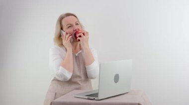 housewife in beige apron at home white background talking to friends on phone eating red apple and watching movie series on laptop carefree life calmness at home Comfort of appeasement gossip problem