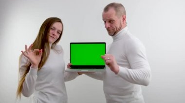 man and girl stand on white background in hands of laptop with green blank screen show gesture with little finger and forefinger goat magical sign protects from evil place for advertising satisfied