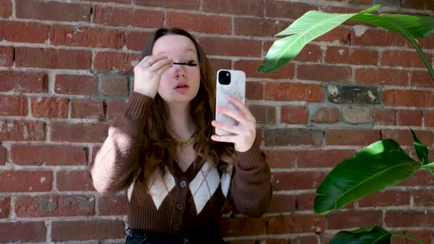レンガの壁の絵を背景に美しい十代の女の子彼女のまつ毛は 携帯電話の花のポットの反射に見えます黒マスカラブラウンブラウスとともにRhombus現代的なファッショナブルな衣装は行く準備をしています — ストック動画