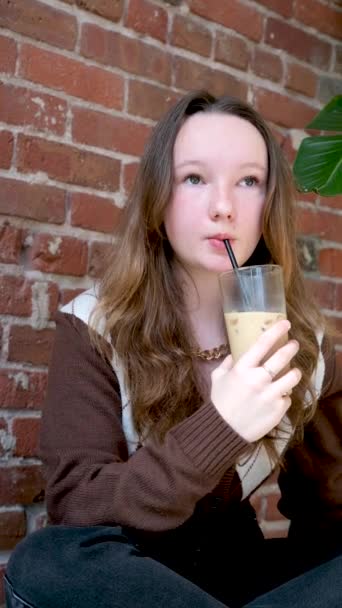 Girl Brown Vest Sits Brick Background Drinks Iced Coffee Cold — Stock Video