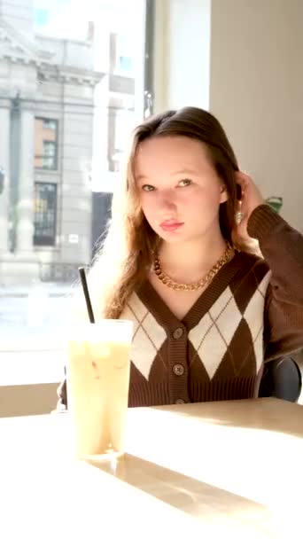 在咖啡店橱窗后面可爱的少女把头发直放在茶几上 加牛奶和冰块的黑管 用来喝褐色外套和菱形的漂亮脸蛋 — 图库视频影像
