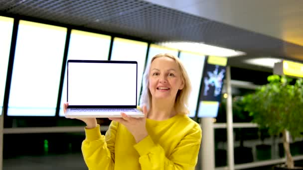 空港の美しい中年女性は彼女の手の中に立っていますノートパソコン広告テキストのための白いモニタースペース笑顔ショーダンス明るい服のスコアボード — ストック動画