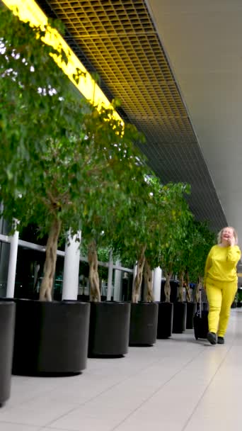 明るいスーツ姿の空港の女性が家に帰りました 車でスーツケースを担いで携帯電話で笑顔で歩いています アムステルダムの行美しい空港に植えられた荷物の木が残りの部分に飛ぶ — ストック動画