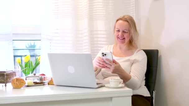 友人と電話で話す女性ノートパソコンの朝食の横にある友人とオンラインでチャットする白いカーテンふっくらブロンドの女性 — ストック動画