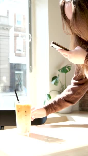 一个有着棕色卷发的少女在阳光下 在透明的玻璃杯中喝杯加冰的咖啡 带着背光视频博客去一家青年咖啡馆拍摄一个关于垂直拍摄食物的视频 — 图库视频影像